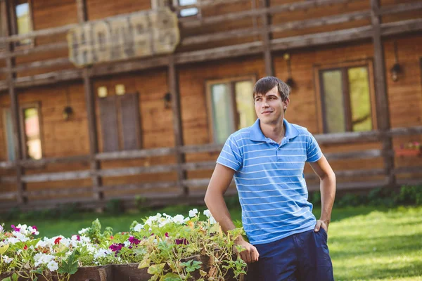 Молодой человек в голубой рубашке и синие шорты возле Телеги декоративные деревянные с цветами на фоне деревенском стиле. Улыбаясь, глядя в сторону — стоковое фото
